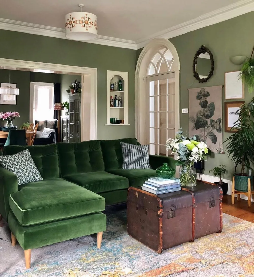 Moody green living room with velvet sectional sofa, botanical artwork, and vintage wooden trunk