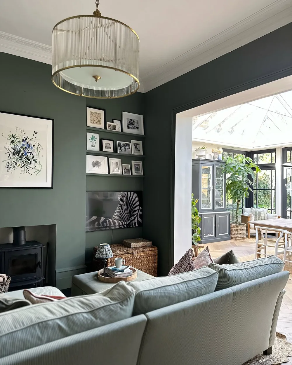 Dark green living room with soft seating, framed artwork, and a bright glass conservatory filled with plants