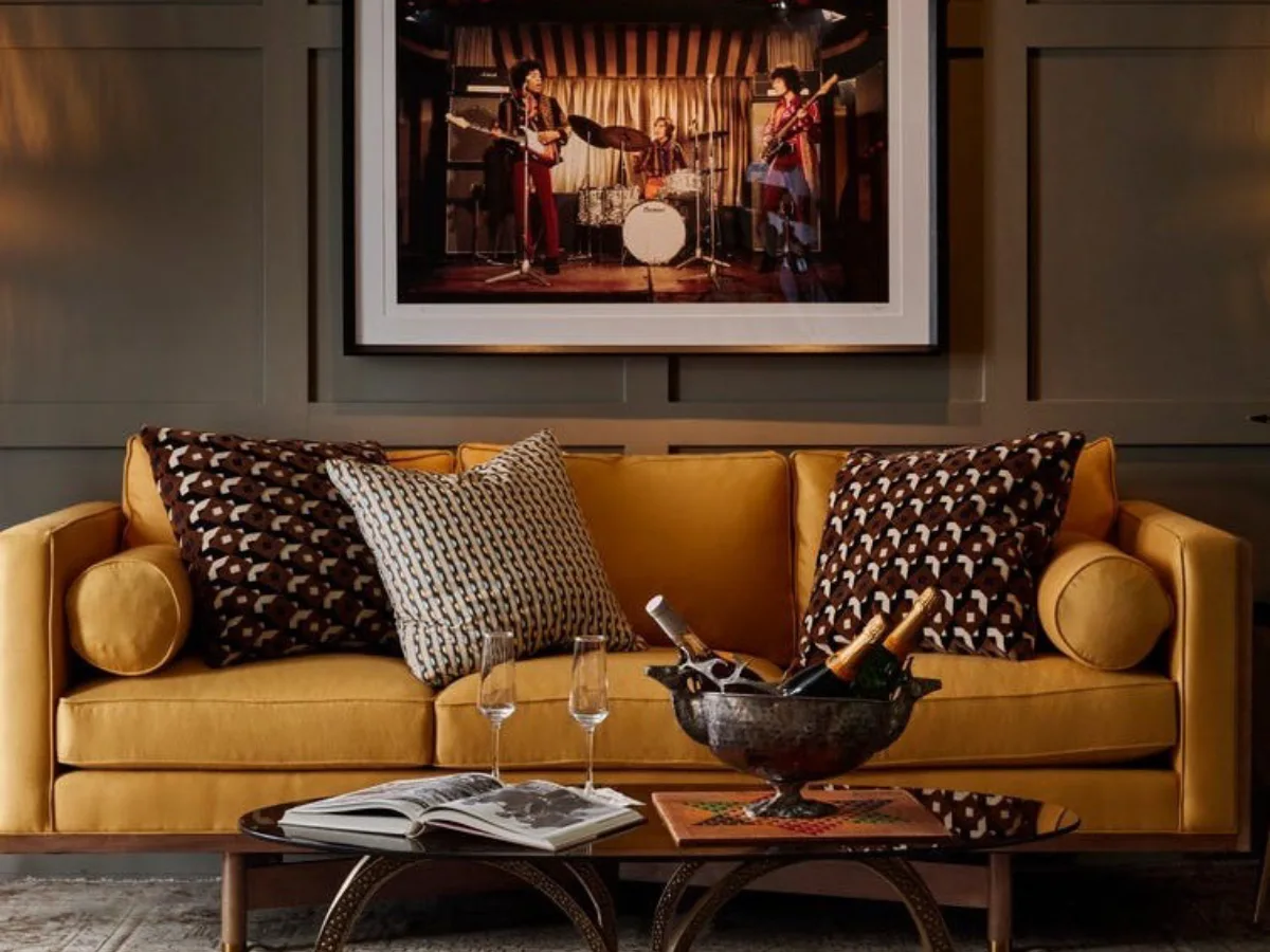 Warm living room with a mustard sofa, gray paneled wall, patterned brown pillows, framed artwork, and a sculptural wooden coffee table.