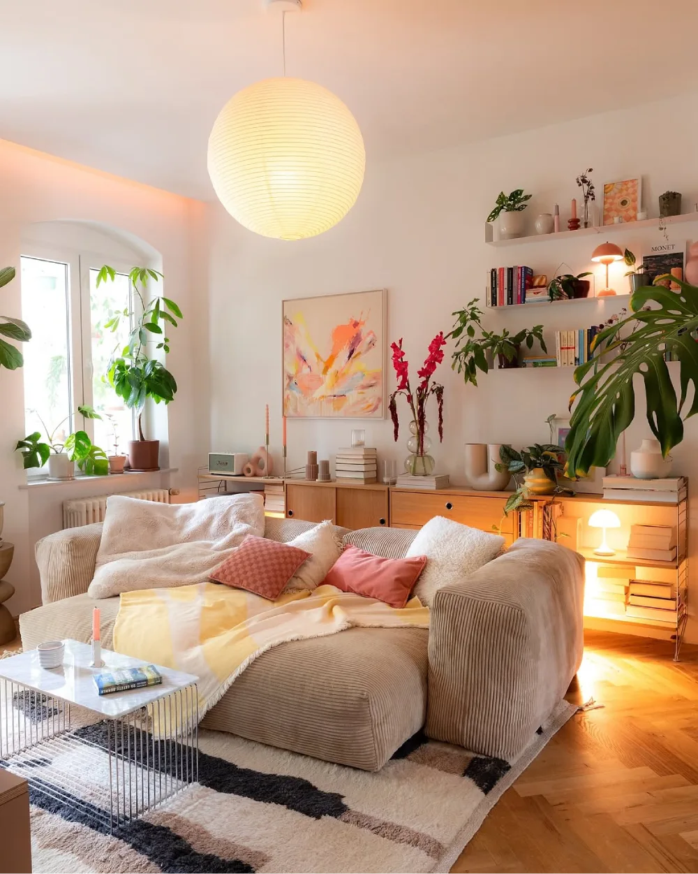 Cozy neutral living room with soft sofa, plants, open shelves, and warm lighting