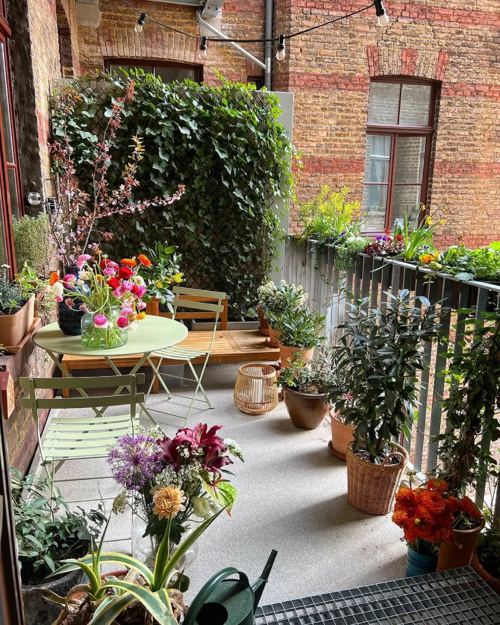 flower-filled balcony with bistro table, green chairs, potted plants, and brick walls