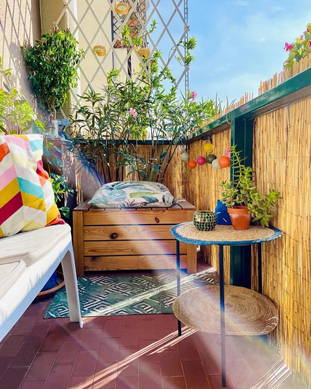sunny balcony with wooden storage bench, bamboo privacy screen, plants, and colorful cushions