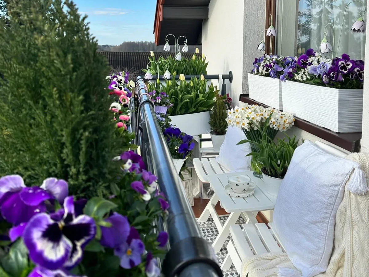 Cozy balcony with white seating, colorful pansies, potted spring flowers, and a small table overlooking a garden.