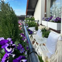 Cozy balcony with white seating, colorful pansies, potted spring flowers, and a small table overlooking a garden.