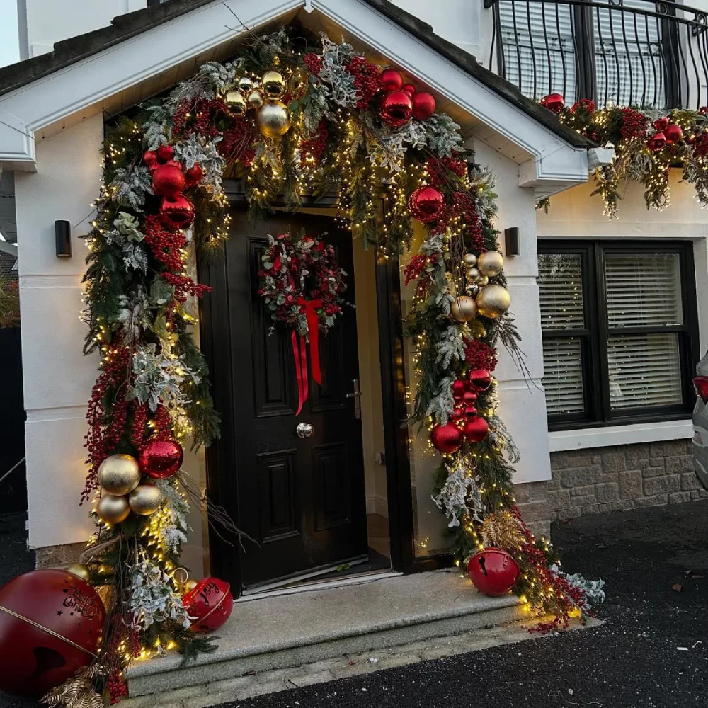 Front door decorated with lush red and gold Christmas garland, ornaments, berries, and warm lights