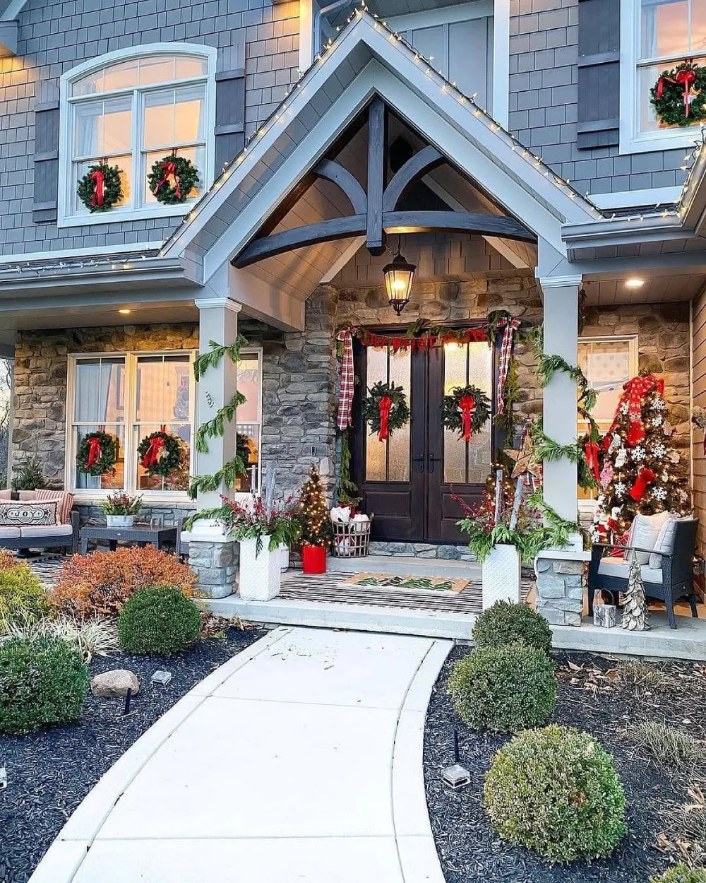 Stone-front porch decorated with evergreen garlands, red bows, wreaths, and Christmas trees