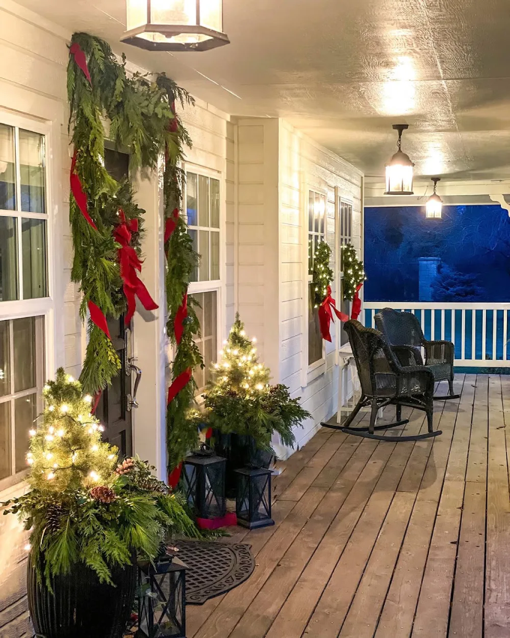 Long covered porch decorated with evergreen garlands, red ribbons, small Christmas trees, and warm lights
