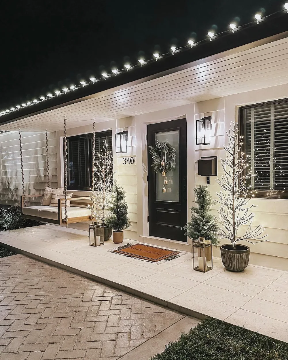 Modern front porch decorated with white Christmas lights, small evergreen trees, and a wreath
