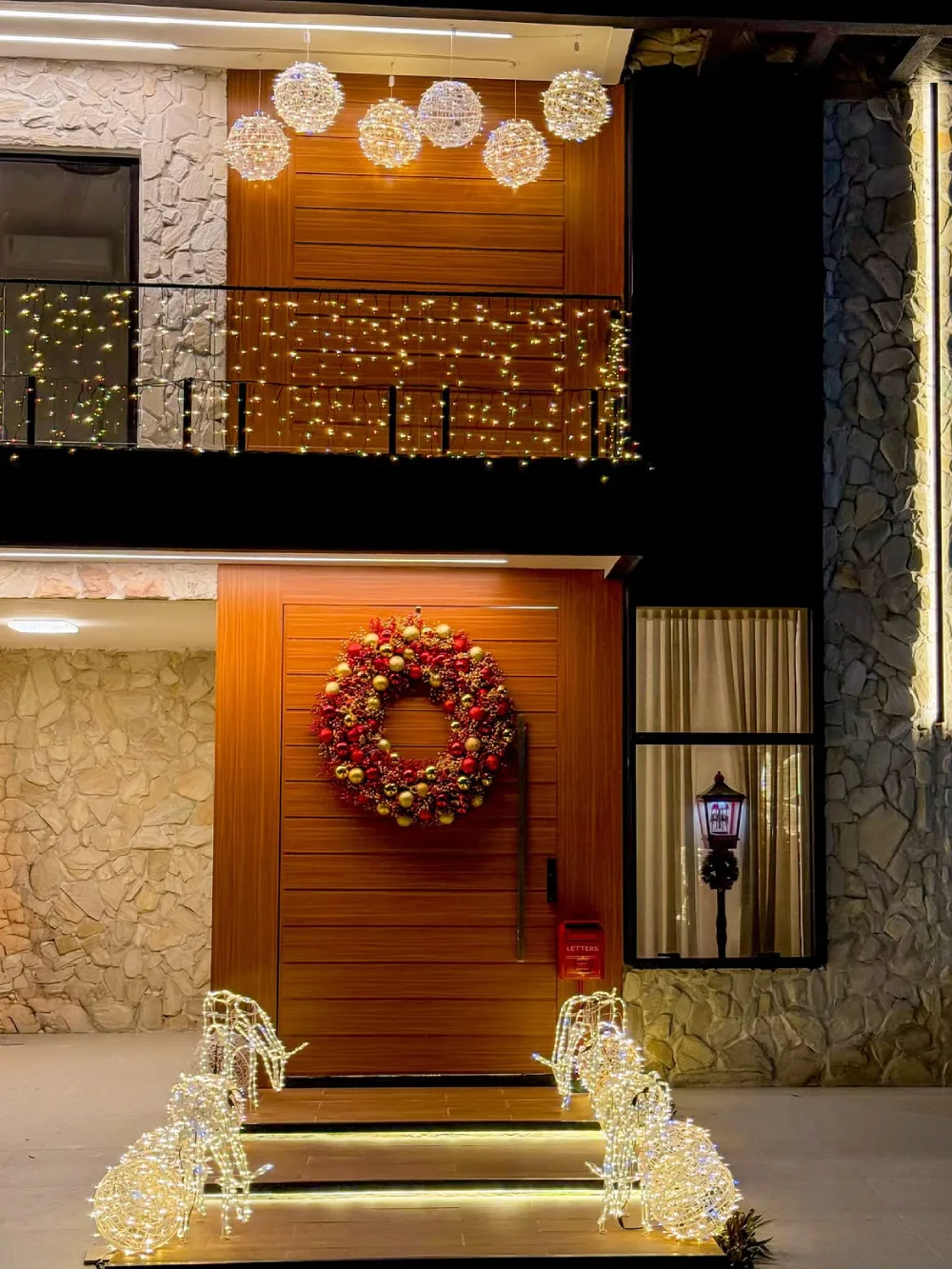 Large modern stone home decorated with warm white Christmas lights and a glowing wreath at the entrance