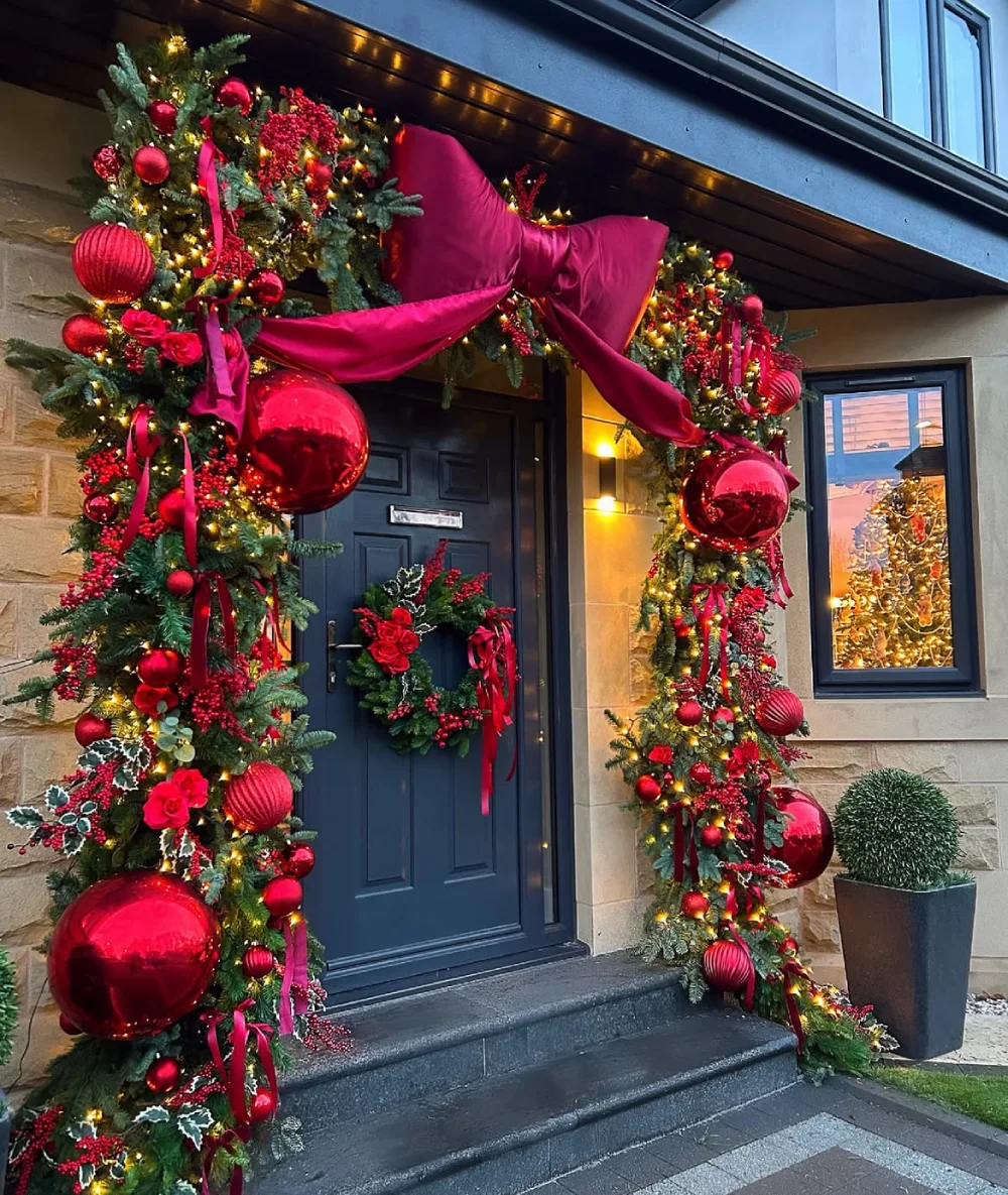 Dark front door framed with Christmas garland, large magenta bow, red ornaments, and warm lights