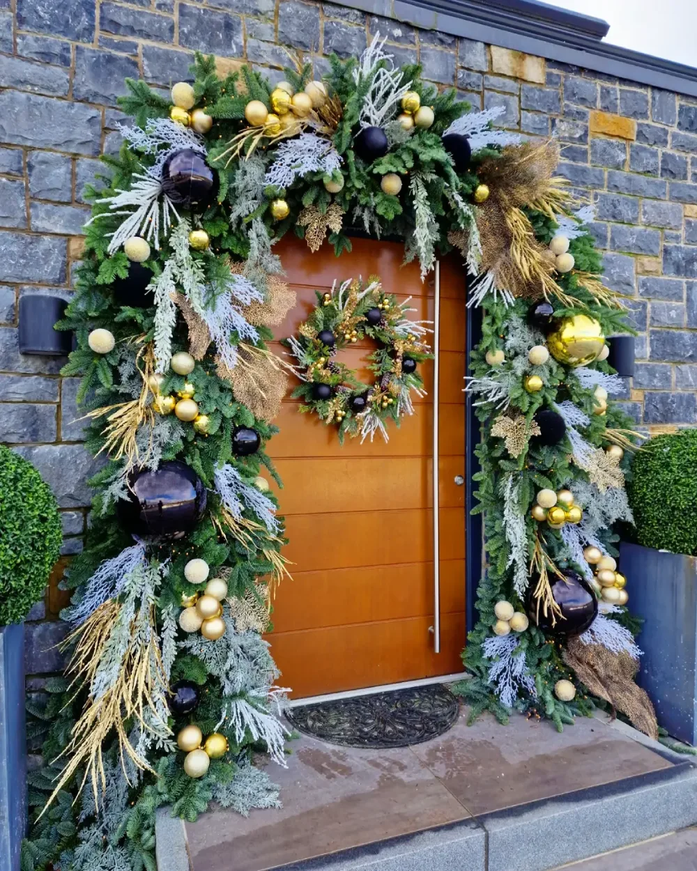 Front door framed with lush Christmas garland, gold ornaments, black accents, and warm lights