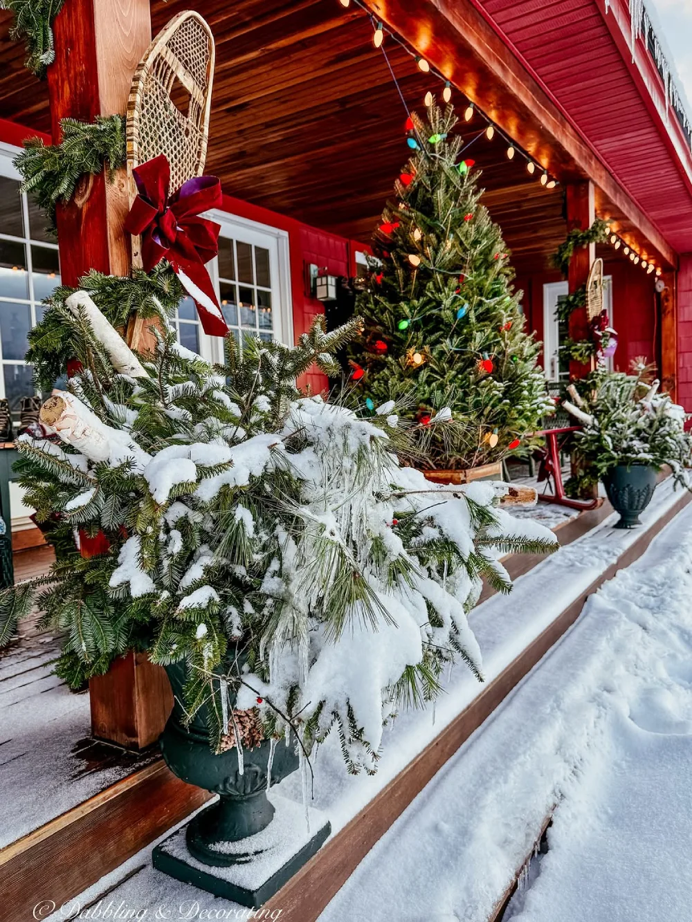 Snow-covered red porch decorated with a Christmas tree, evergreen garlands, and colorful lights
